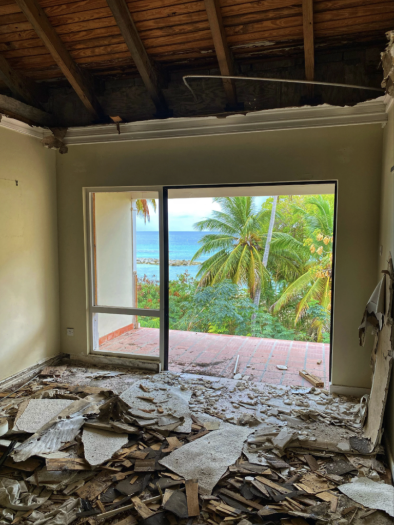 A room under renovation with debris on the floor, an exposed ceiling, and a large glass door. The view outside shows palm trees and a glimpse of the ocean under a blue sky.