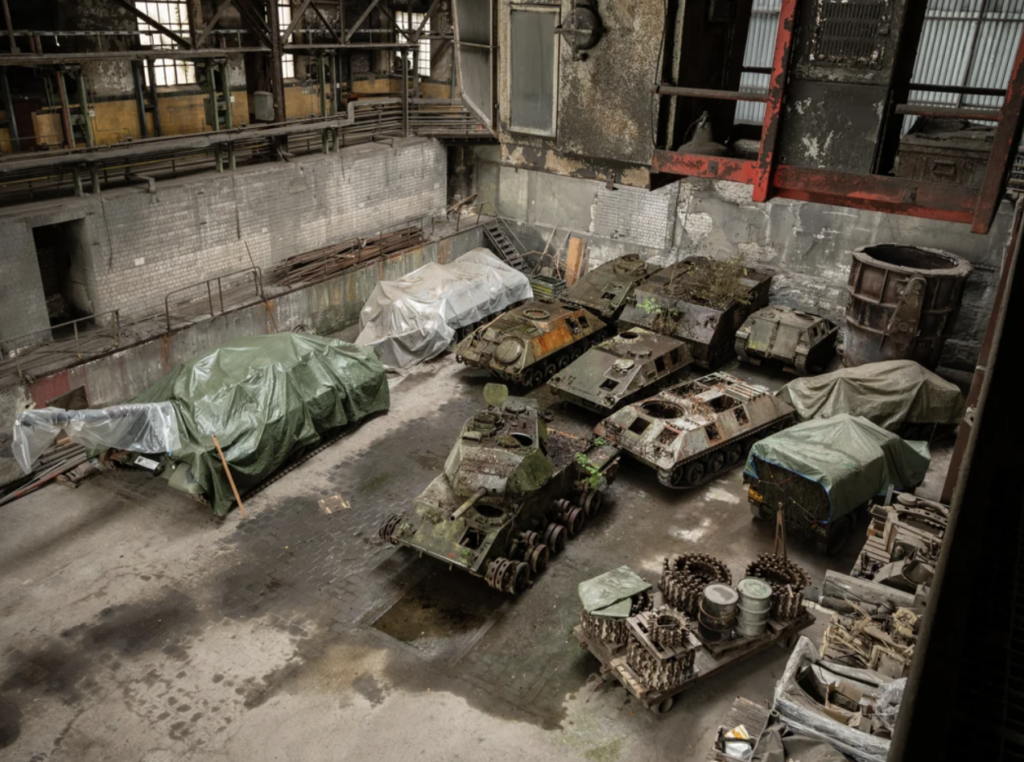 A photograph shows an old industrial warehouse with several abandoned, rusting military tanks. Some are partially covered with tarps. The setting is dilapidated, with debris and metal parts scattered around.