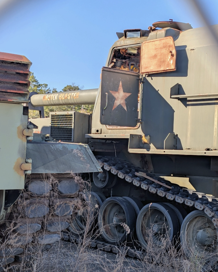 A weathered military tank with a prominent star emblem and the words "Quicker Blaster" painted on its barrel. The vehicle is set on a grassy field under a clear blue sky.