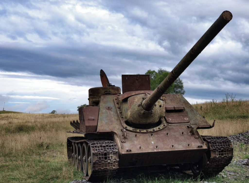 An old, rusted military tank sits abandoned in a grassy field under a cloudy sky. The tank's turret is aimed forward, and its tracks are partially visible. The scene conveys a sense of decay and history.