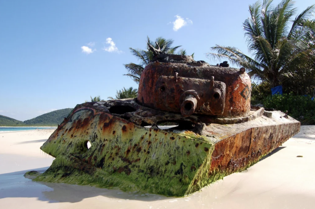 Rusty tank half-buried in sand on a beach, showing signs of decay with green moss at its base. Surrounded by palm trees, clear blue sky, and mountains in the distance.