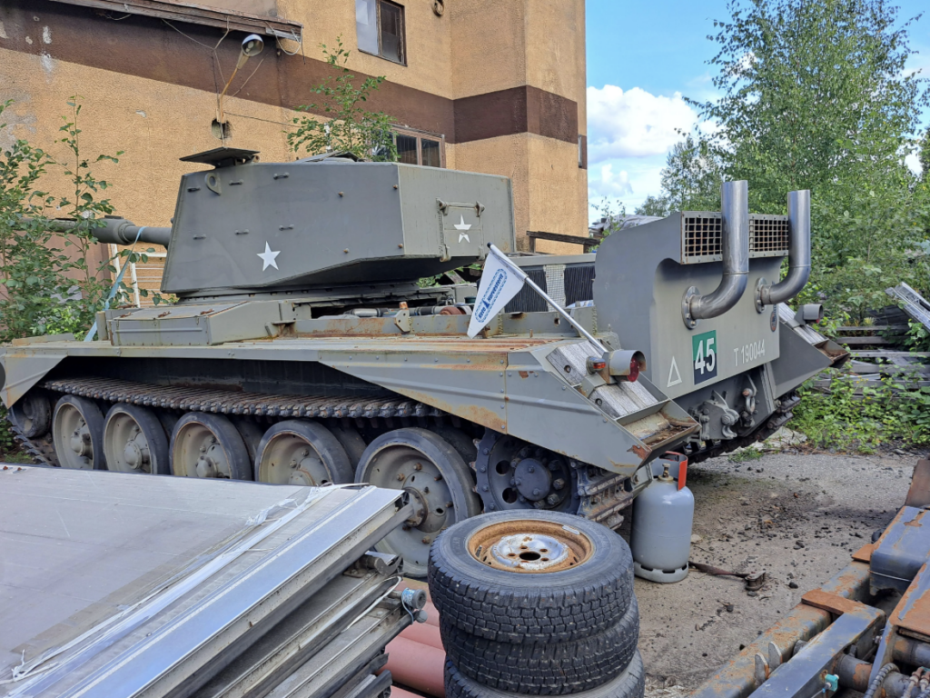 A vintage military tank with a number "45" and insignias is parked outdoors. It's surrounded by some greenery, discarded tires, and metal sheets. The setting appears to be an industrial or storage area.