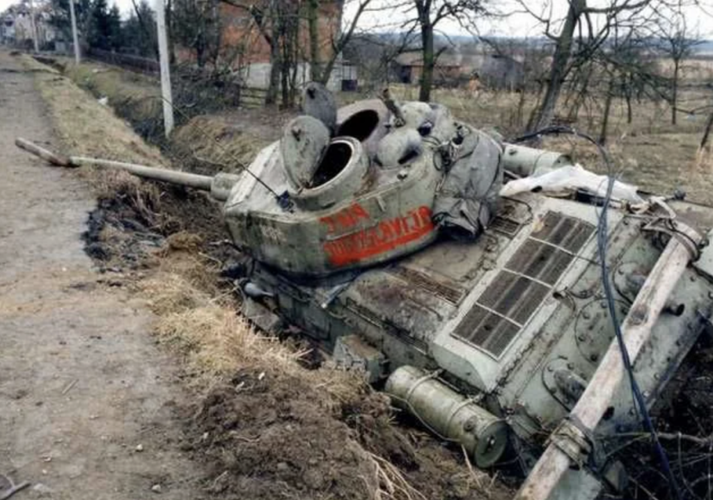 A heavily damaged, overturned military tank lies in a muddy ditch beside a dirt road. The tank appears weathered and rusted, with part of its turret open. Bare trees and a brick building are visible in the background.