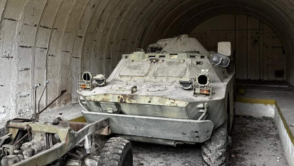 A dusty, abandoned military armored vehicle is stationed in a dimly lit, arched concrete shelter. The vehicle appears weathered, with visible rust and dirt, and is positioned near an old, rusty trailer. Debris is scattered on the ground.