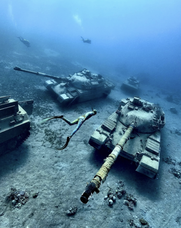 Divers explore submerged tanks on a seafloor, covered in coral and marine growth. The scene is illuminated by natural sunlight filtering through the water, and the tanks lie in various positions, creating an intriguing underwater landscape.