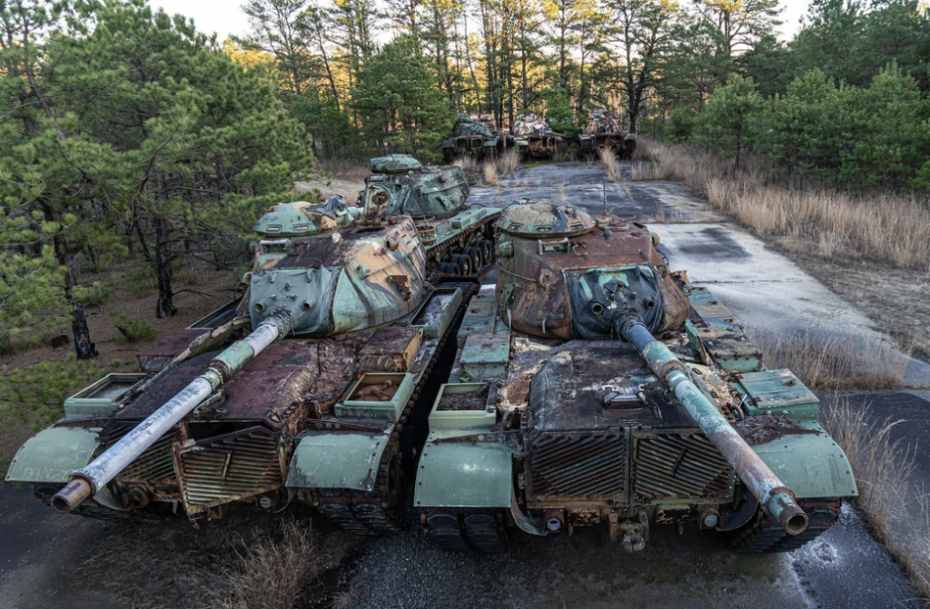 Rusty, abandoned tanks lined up on a cracked, overgrown road surrounded by trees. The tanks are weathered, with peeling paint and significant decay, set under the dim light of a cloudy sky.