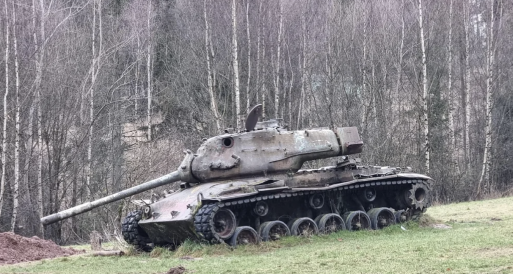 A rusted, abandoned tank sits on a patch of grass in a forested area. Bare trees with thin trunks form the background, suggesting a winter or early spring setting. The tank's paint is faded, and it appears weathered.