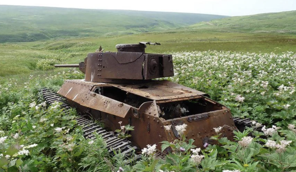 A rusted, abandoned tank sits in a field of wildflowers amidst rolling green hills. The tank is overgrown with vegetation, blending into the lush landscape under a cloudy sky, evoking a sense of historical decay and nature's reclamation.