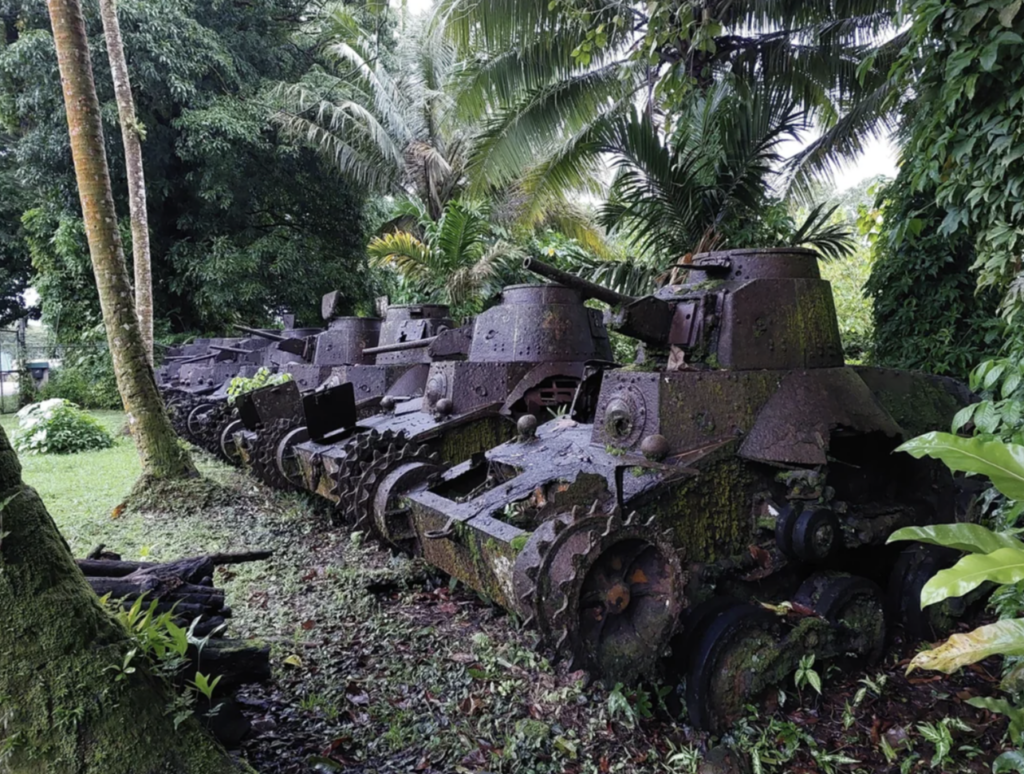 Several rusted World War II-era tanks are partially covered in moss and vegetation, sitting in a tropical forest setting. Tall palm trees and dense greenery surround the tanks, suggesting nature's reclamation of the area.