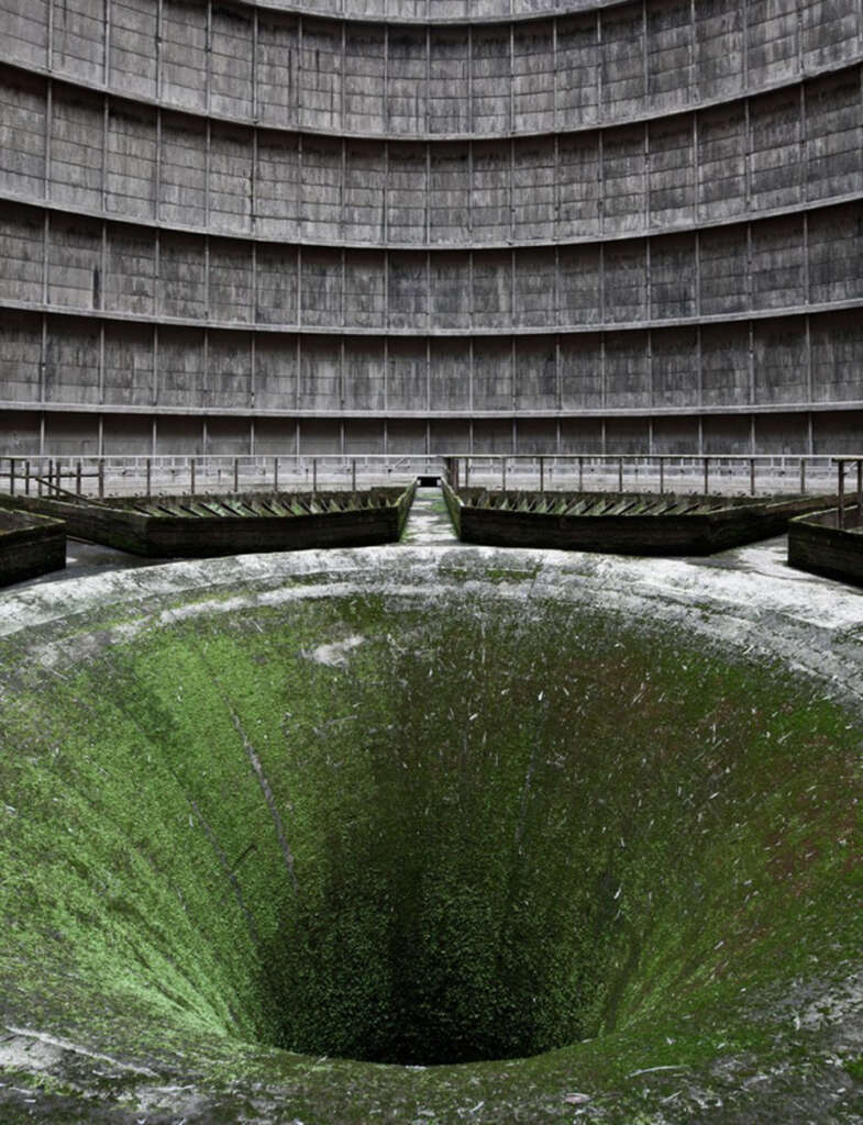 Interior of an abandoned cooling tower with a large, moss-covered funnel-like structure at the base. The decaying walls rise in concentric circles, creating a dramatic perspective showing the tower's height.