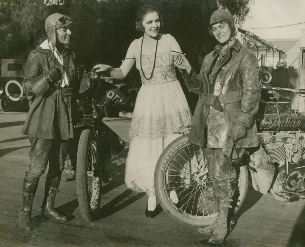 Three people stand outdoors near vintage motorcycles. Two are dressed in early 20th-century motorcycle gear, while the third is in a light dress with a long necklace. They smile and pose together, with trees and cars visible in the background.