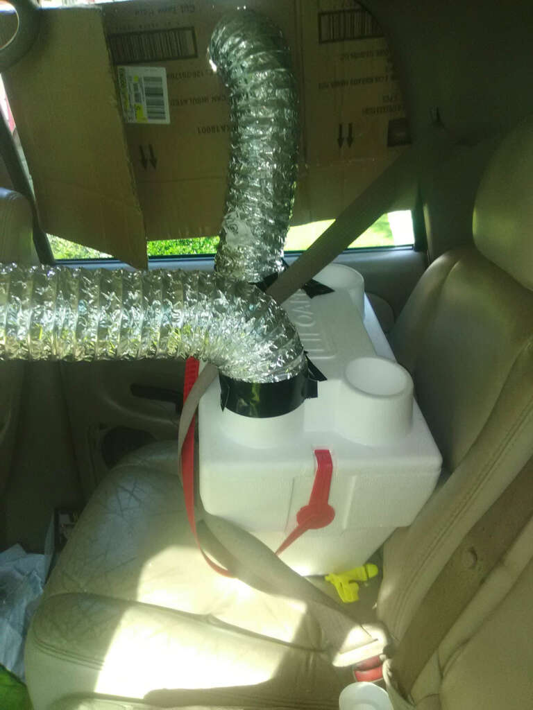 A homemade air conditioning unit sits on a car seat. It includes a large white cooler with metal duct tubes attached. The cooler is secured with a seatbelt and surrounded by a cardboard box with additional ductwork leading to the window.