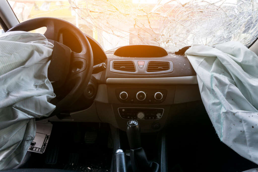 A car interior with deployed airbags from the steering wheel and passenger side, and a shattered windshield. The dashboard features various controls, and the sunlight is visible through the damaged glass.