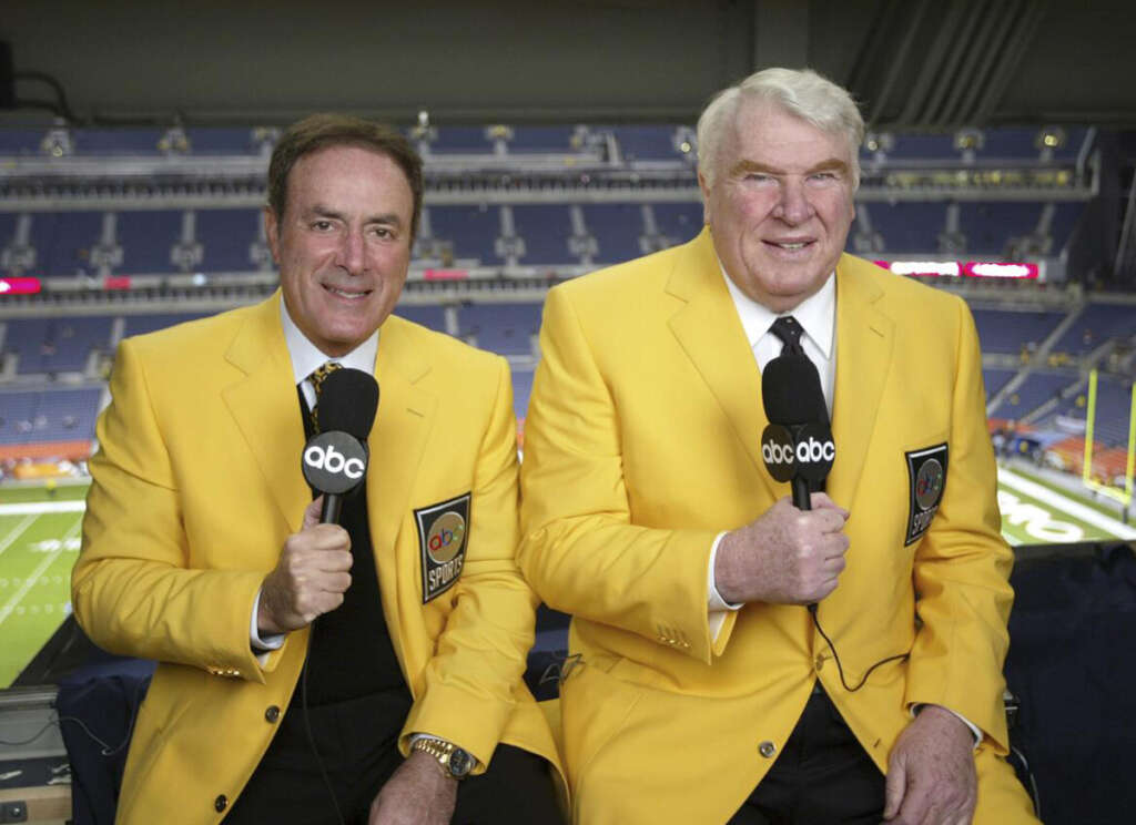 Two sports commentators wearing bright yellow jackets sit in a booth at a stadium. They are holding ABC microphones and smiling. The football field is visible in the background through the window.