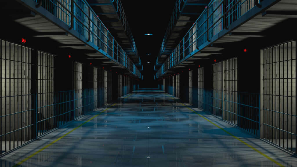 Dimly lit prison corridor with metal-barred cells on both sides. The shiny floor reflects the blue-toned overhead lighting. Red lights are above some cell doors, and metal walkways are visible on the upper level.