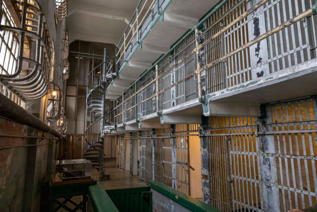 Interior view of a vintage prison cell block with multiple tiers. Iron bars line the cells, and a spiral staircase connects the floors. The aged metal and worn paint give the ambiance a historic and weathered appearance.