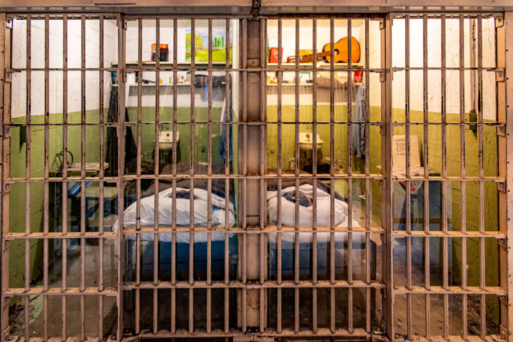 A prison cell with a barred door. Inside, two beds with blue mattresses are visible. The walls are painted green and white. On the back wall, shelves hold various items, including books and a guitar. A sink and toilet are also present.