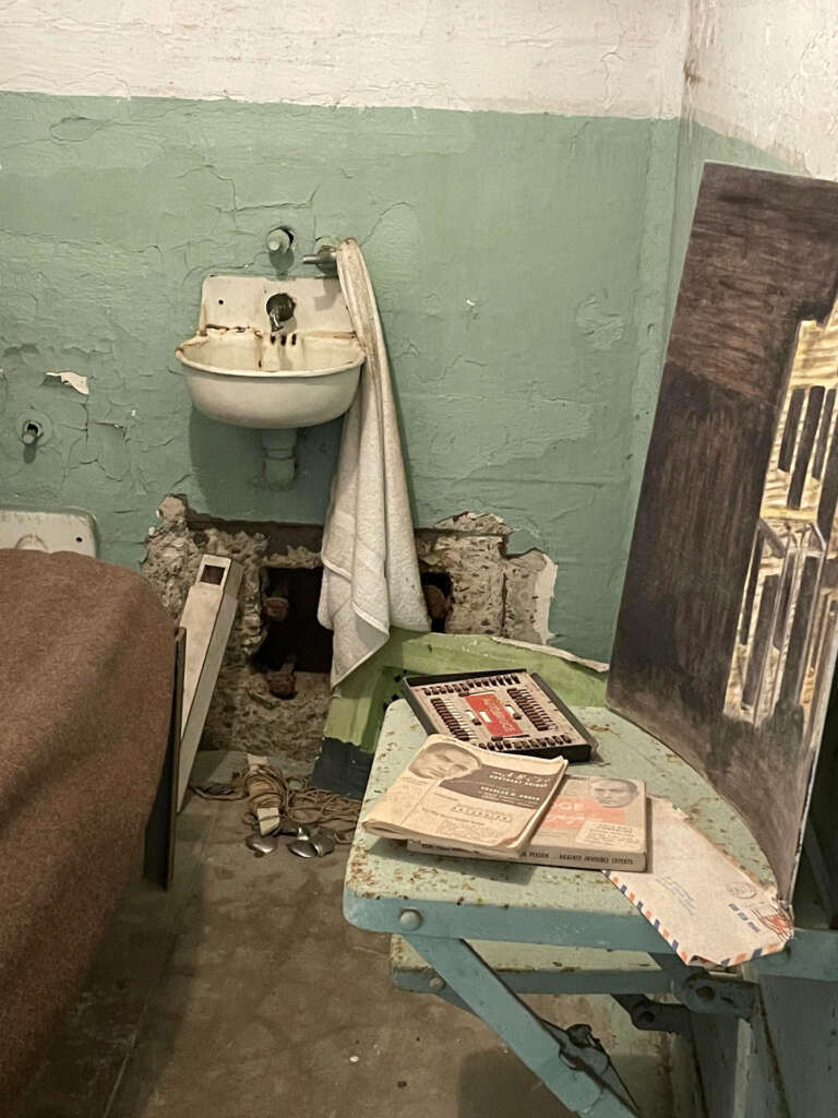 A small prison cell with a bed, a sink with a towel hanging, and a green chair. On the chair are books and a notepad. The walls are worn, and a small window allows light into the dim room.