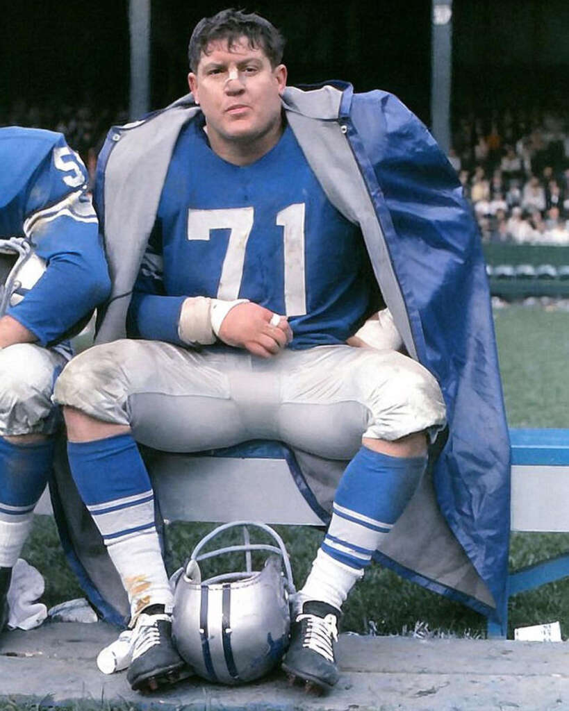 A football player wearing a blue jersey with the number 71 sits on a bench. He has a coat draped over his shoulders and a silver helmet at his feet. The background shows a football field and spectators.