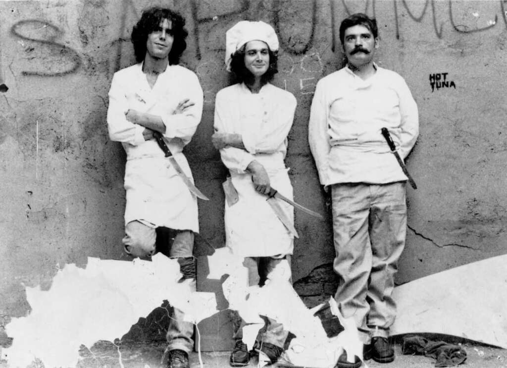 Three people dressed as chefs stand against a graffiti-covered wall. The person on the left holds a knife, the one in the middle wears a chef's hat, and the one on the right has a knife tucked in their apron. The photo appears aged and torn.