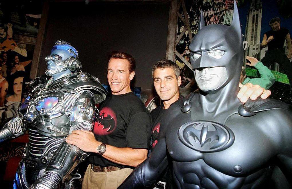 Two men stand between life-sized models of a robotic figure and a Batman figure. The men are smiling and pose with their arms around the models, in a room filled with posters and memorabilia.