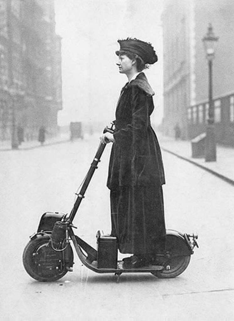 A woman in early 20th-century attire stands on a vintage scooter on an empty city street. She's dressed in a long coat and hat, looking forward. The scene is in black and white, depicting a historical urban setting.