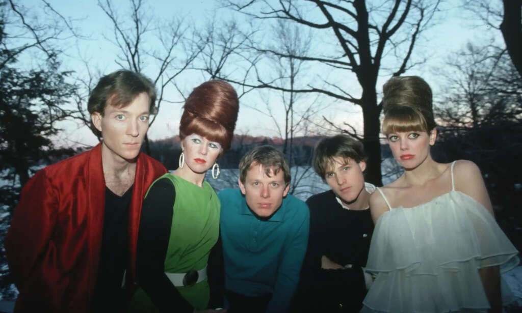 Five people are posing outdoors during twilight. Two women have beehive hairstyles and wear eye-catching outfits, while three men are dressed more casually. Bare trees and a fading sunset create a serene backdrop.