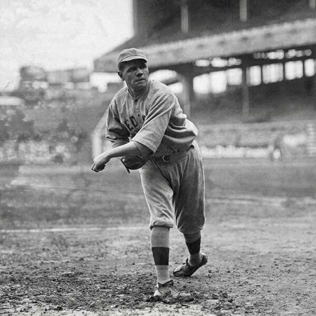 A vintage photo of a baseball player in mid-pitch on a dirt field. He's wearing a uniform with "RED" partially visible on the chest and a cap. The background shows a large, partially filled stadium with spectators.