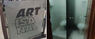 Split image: Left shows a glass gate with the bold text "ARTISANAL." Right shows a bathroom with two adjacent stalls; the frosted glass partially conceals toilets, providing minimal privacy.