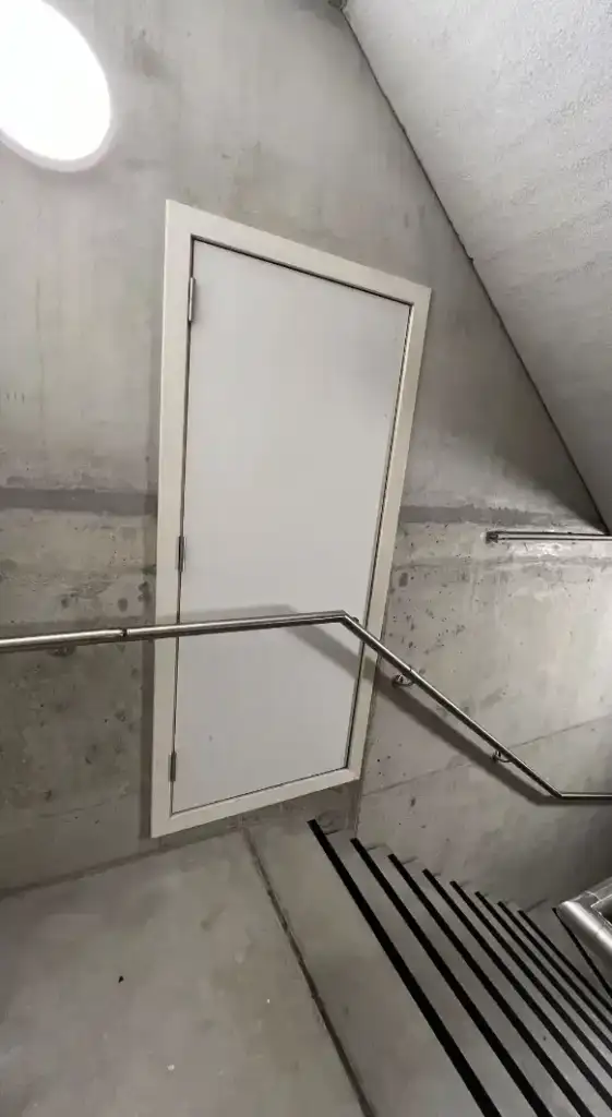 A white door positioned midway on a concrete wall in a stairwell, with a metal handrail leading upward. The stark and minimalist surroundings emphasize the unusual placement of the door.