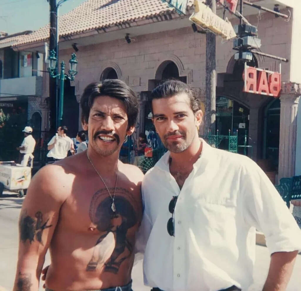 Two men pose together outdoors. The man on the left is shirtless with tattoos and a necklace, and has a mustache and styled hair. The man on the right wears a white button-up shirt and has short hair. A building with a bar sign is in the background.