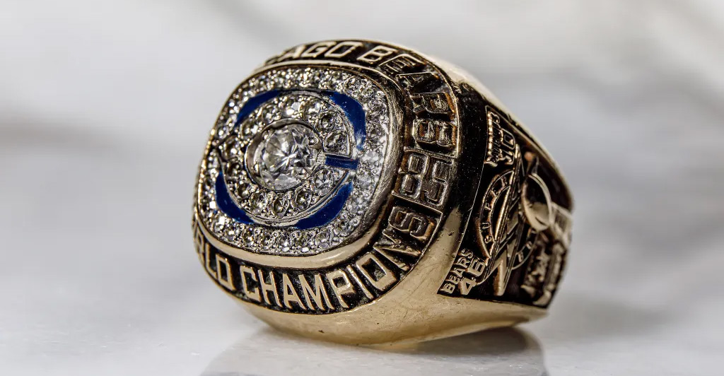 Close-up of a gold championship ring with a large diamond center, surrounded by smaller diamonds. "Chicago Bears World Champions" is engraved on the ring, and the letter "C" is prominently featured in blue.