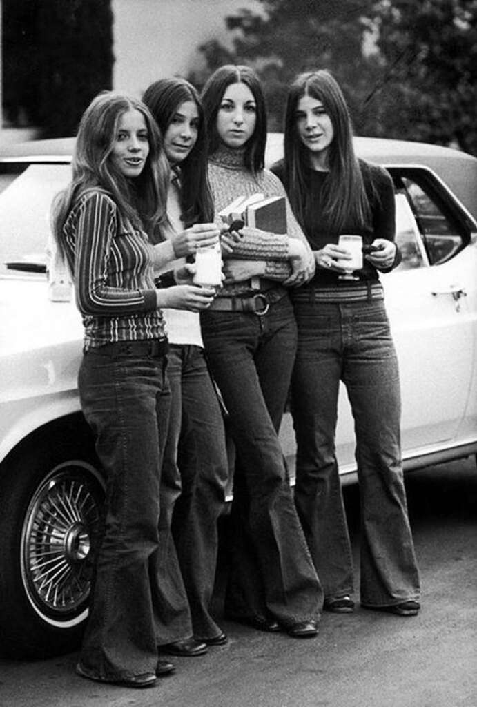 Four women in 1970s fashion stand in front of a car. They wear flared jeans and long-sleeve tops, holding drinks. The scene has a vintage feel, with trees in the background and one person carrying a book.