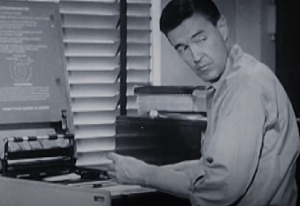 A man in a long-sleeve shirt sits at a desk, looking toward a typewriter. The room has closed blinds and several books are stacked on the desk. The image is in black and white.