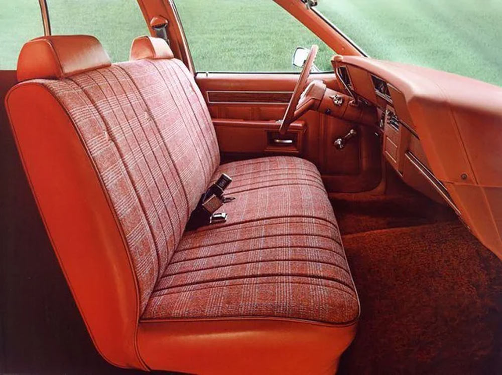 Vintage car interior featuring orange and plaid bench seat, matching door panels, and carpet. Steering wheel and dashboard are up front. Background shows a green outdoor setting.