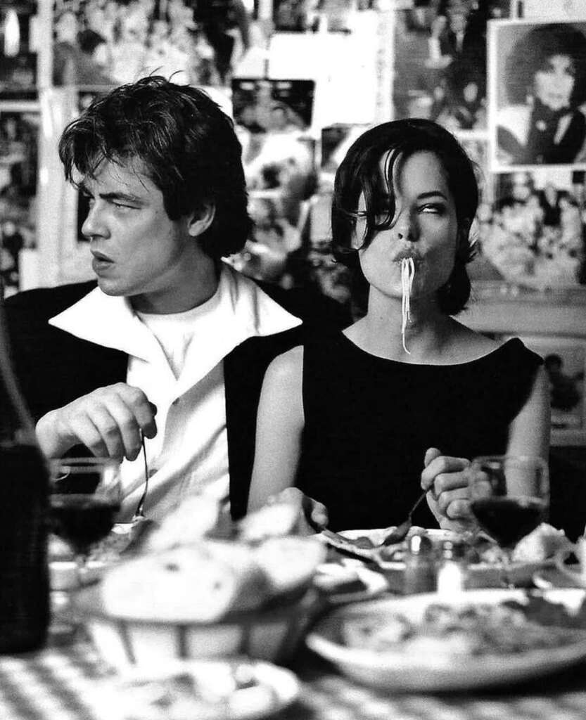 A black and white photo of two people dining at a table. The man is looking to his left with his mouth slightly open, while the woman is eating spaghetti with a playful expression. The table is set with wine glasses and bread. Various photos cover the wall.