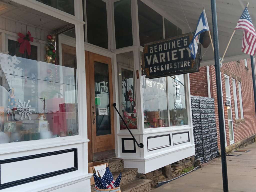 Storefront of Berdine's Variety, a 5&10¢ store, with vintage decor. The entrance has wooden doors and festive decorations. American and other flags are displayed. The windows show various items and a snowflake design. Red brick walls are visible.