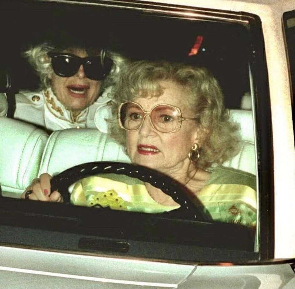 Two elderly women in a car. The driver is wearing large glasses and a green top, gripping the steering wheel. The passenger in the back has curly hair, dark sunglasses, and a white outfit, smiling.