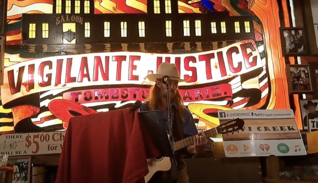 A musician wearing a hat plays an acoustic guitar in front of a vibrant, illuminated backdrop with the words "Vigilante Justice Tombstone Ariz" and various signs and symbols. A tip jar and a red cloth cover part of the stage.