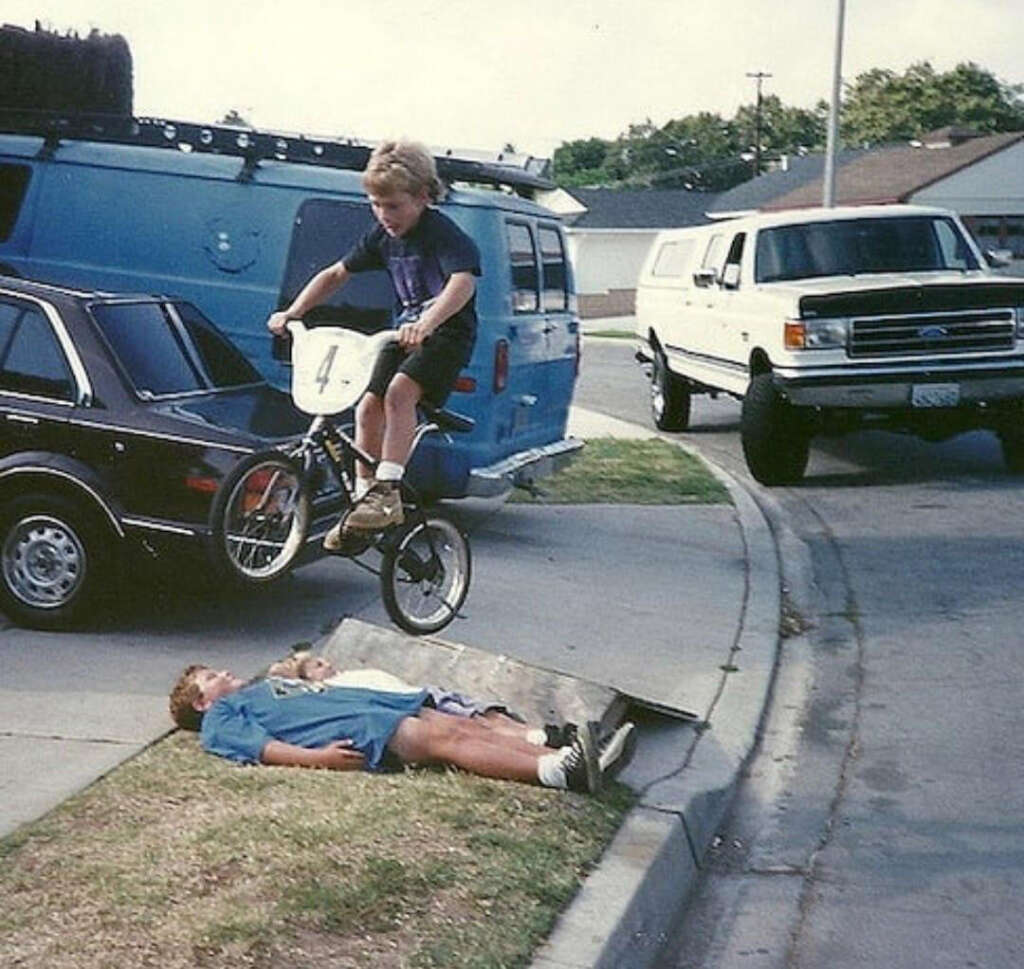 A child on a bicycle jumps over another child lying on the grass next to a curb. The scene is in a suburban neighborhood with two parked vehicles nearby, a blue van and a white truck. The biker is airborne, performing the stunt.