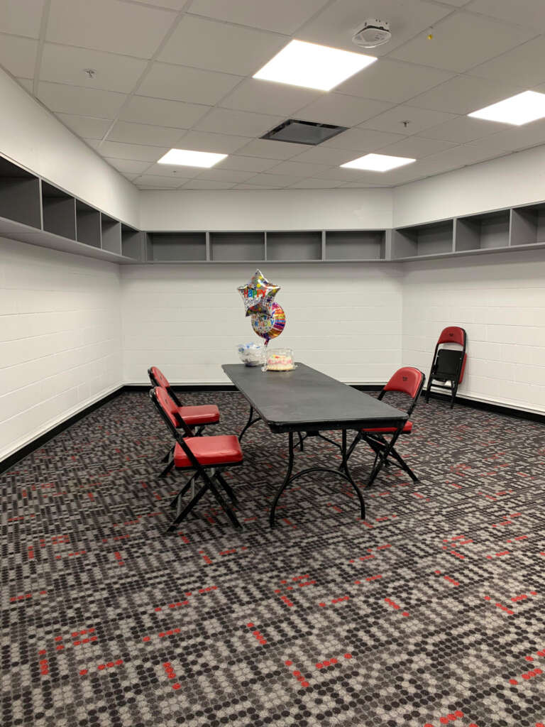 A small, undecorated room with red and black patterned carpet contains a folding table with four red chairs. On the table are balloons and a cake. The room has empty gray shelves and a stack of chairs in the corner.