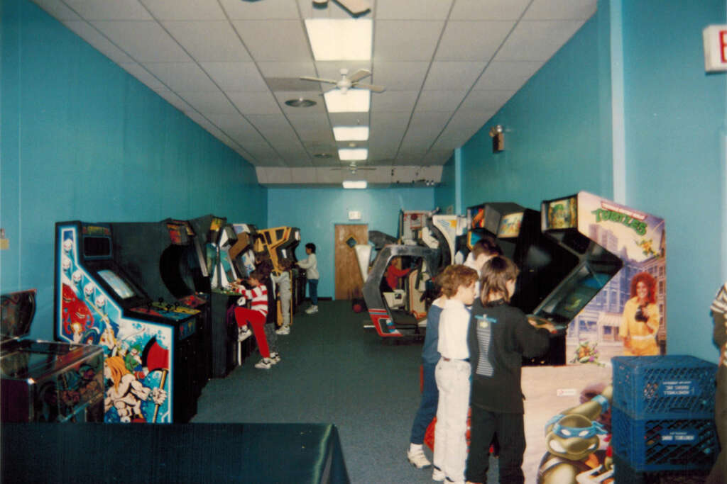 A group of children are playing various arcade games in a retro gaming arcade with blue walls. The room is filled with vintage arcade machines, including pinball and racing games. The setting has a nostalgic, 80s or 90s vibe.