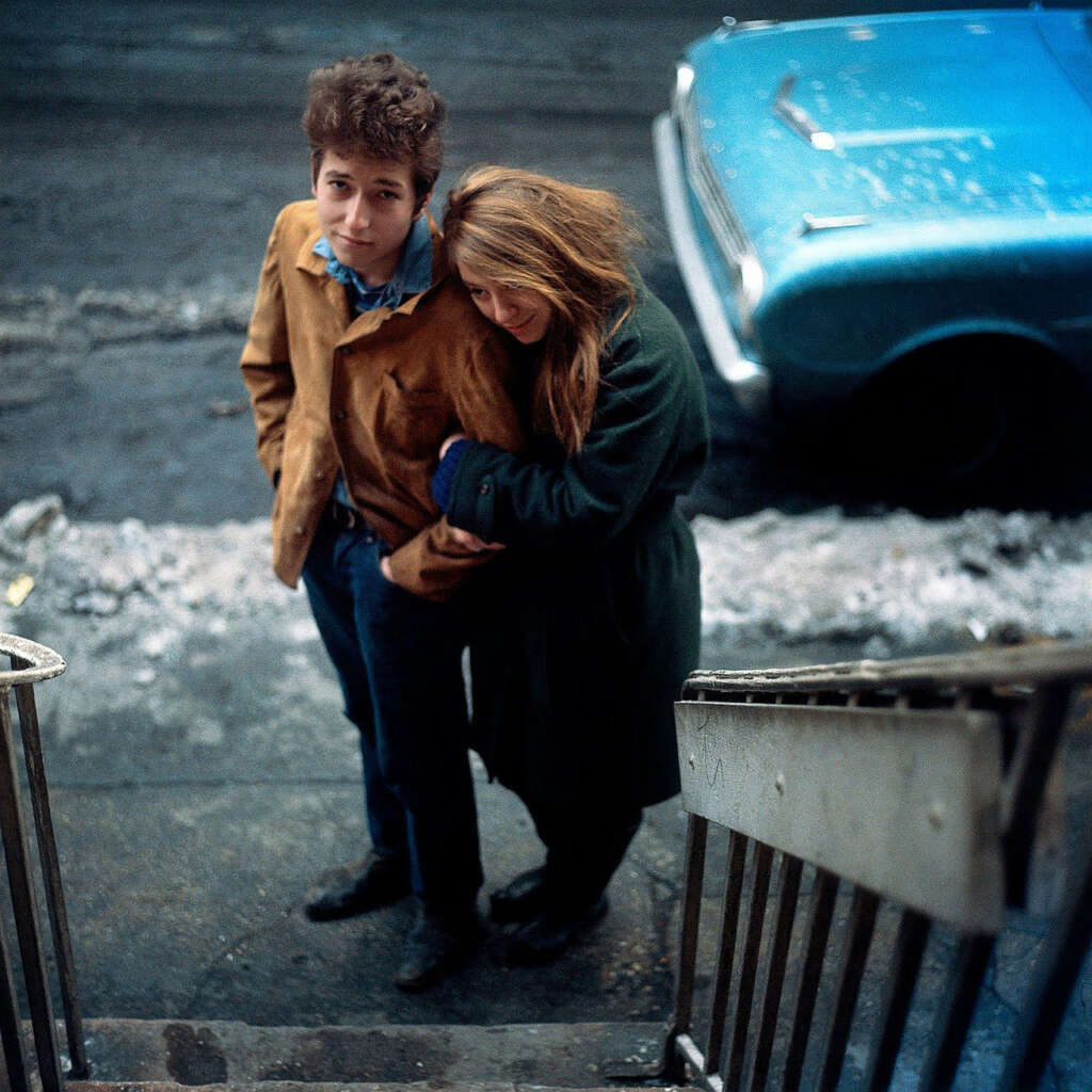 A man and woman stand closely on a city sidewalk, viewed from above. The man wears a brown jacket, the woman is in a dark coat. A blue vintage car is parked nearby. The street has snow along the curb. Steps in the foreground lead down to the sidewalk.