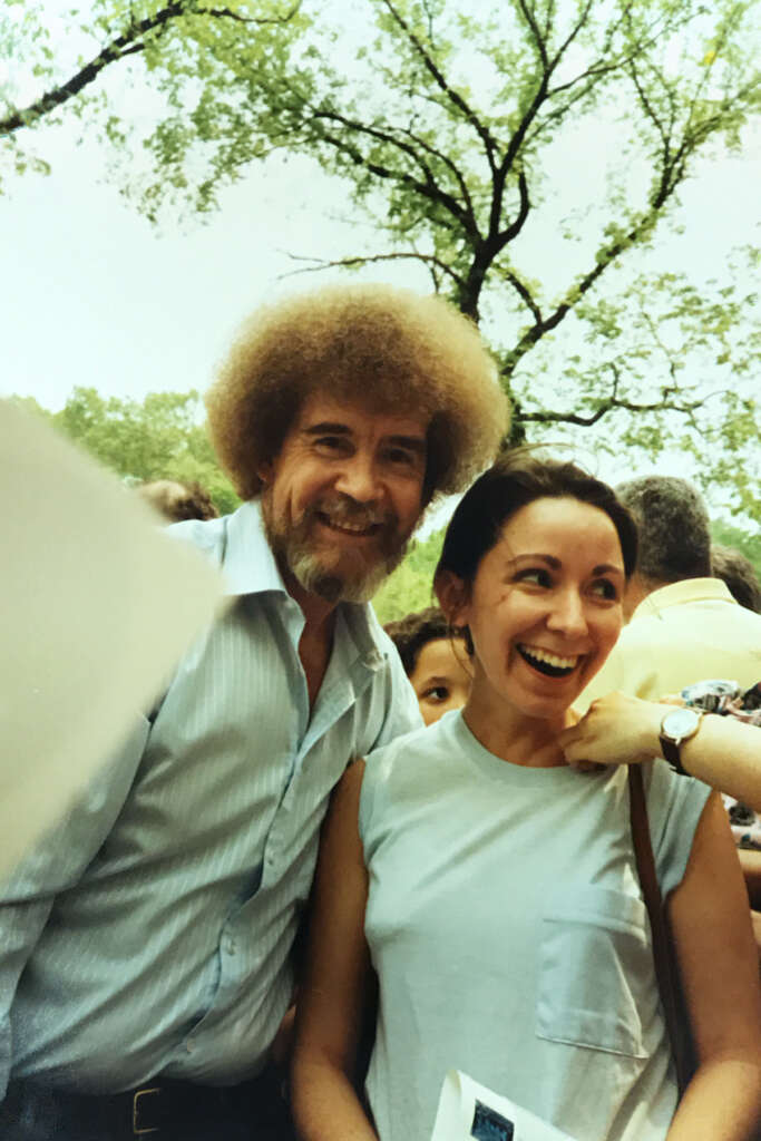 A man with curly hair and a beard stands smiling next to a woman in a sleeveless top. They are outdoors with trees in the background. Other people are partially visible around them.