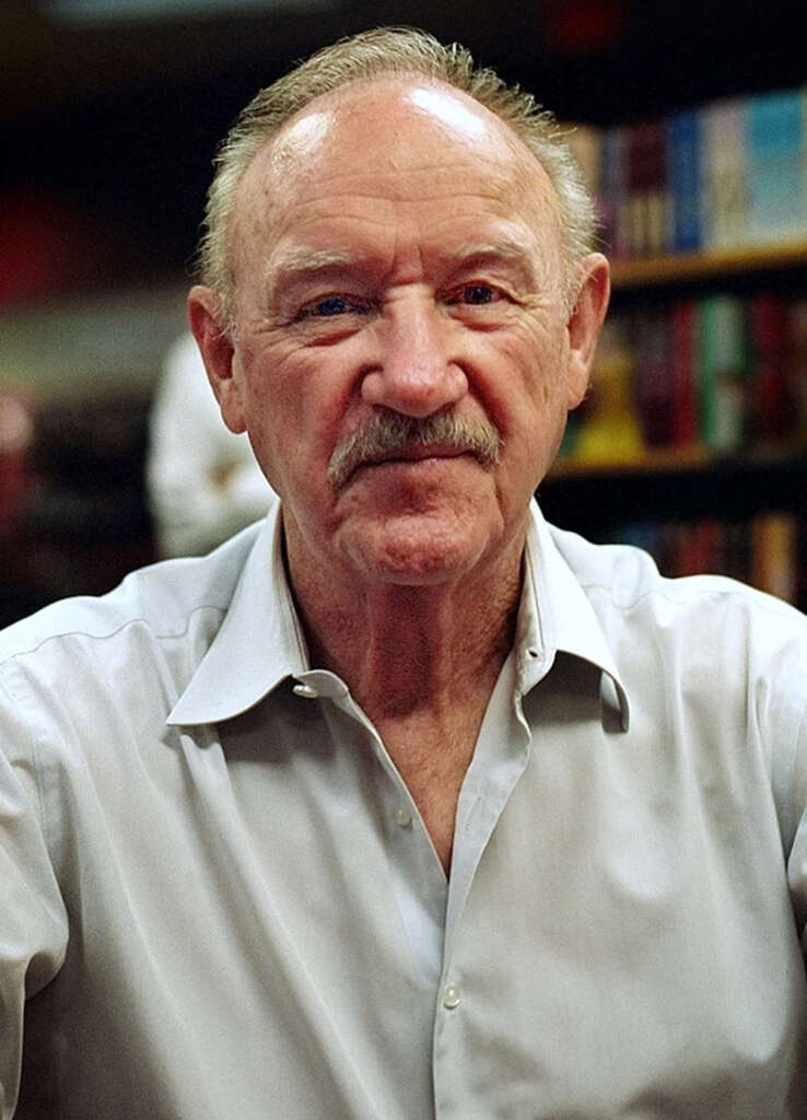 An older man with a mustache and receding hairline is wearing a light gray shirt. He is seated in front of shelves filled with colorful books. The background is slightly blurred, focusing on the man.