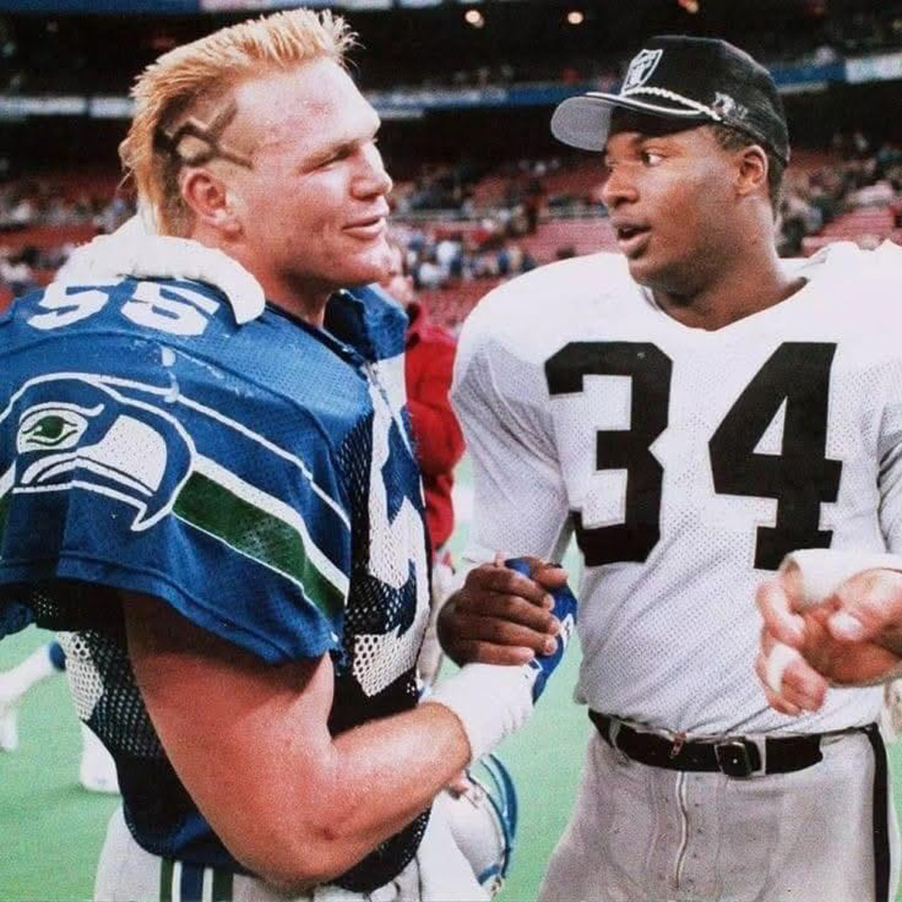 Two American football players stand on a field, shaking hands and smiling. One wears a blue Seattle Seahawks jersey and the other a white jersey with the number 34. They appear to be having a friendly conversation after the game.