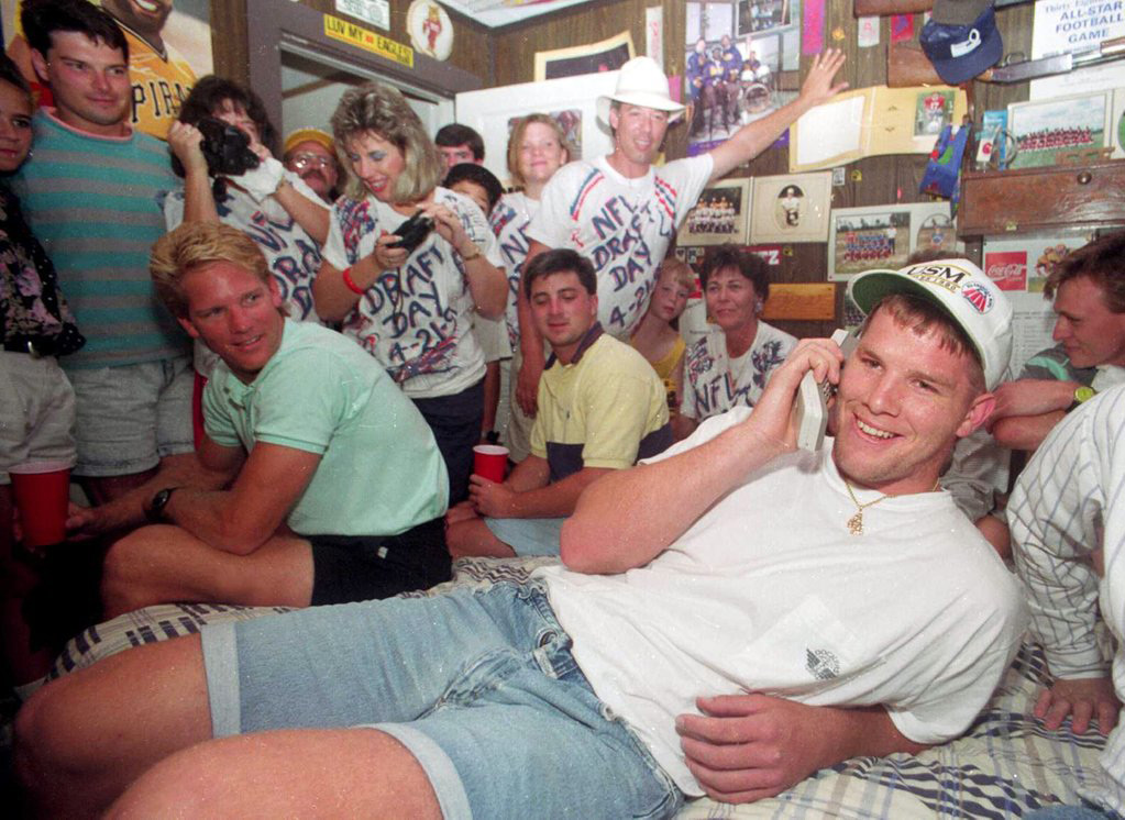 A group of people in a room during an NFL draft party. The central figure, Brett Favre is sitting on a bed, is talking on a cellphone, smiling. Others are gathered around, some wearing "NFL Draft Day" shirts, looking excited and engaged.