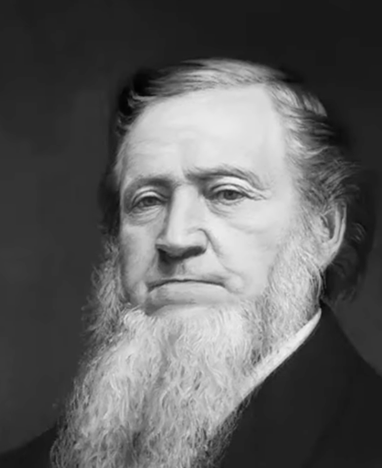 A sepia-toned portrait of an older man with a long, full, white beard and a serious expression. He is wearing a dark suit jacket and a light-colored shirt. The background is plain and dark, emphasizing his features.