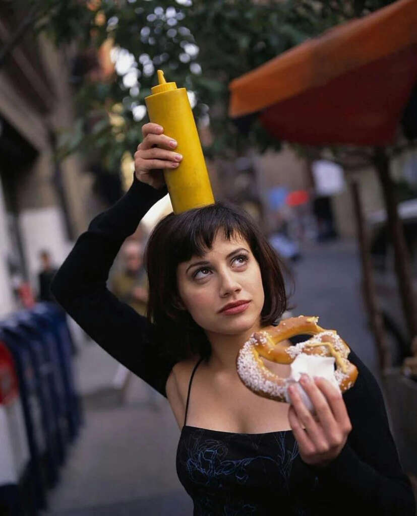 A woman with short dark hair playfully balances a large yellow mustard bottle on her head. She holds a pretzel with mustard in her other hand. The background shows an outdoor urban setting with blurred buildings and trees.