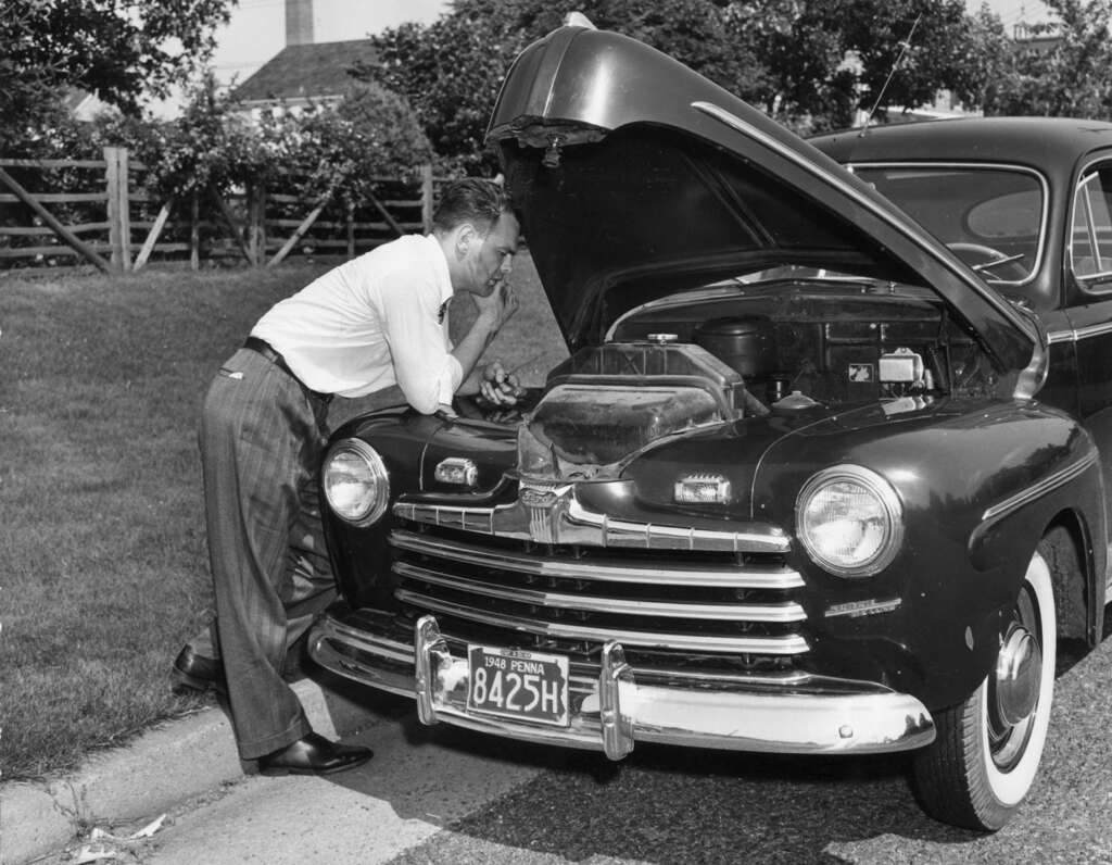 A man in a white shirt and plaid pants leans over the open hood of a classic car, inspecting the engine. The vintage car has a license plate number 8425H. Trees and a house are visible in the background.